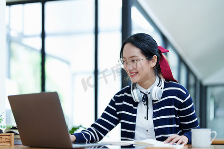 一名亚洲少女使用电脑、戴着耳机、使用笔记本通过图书馆木桌上的视频会议在线学习的肖像