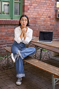 亚洲女孩的全尺寸肖像坐在笔记本电脑上，看起来很惊讶和惊讶，并偷看屏幕。