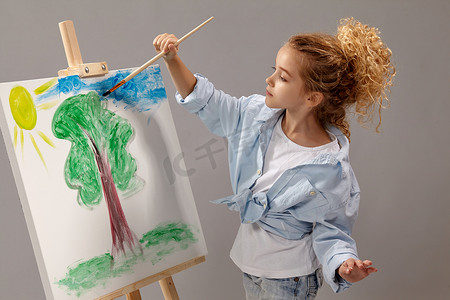 迷人的女学生站在灰色的背景上，用水彩画笔在画架上绘画。