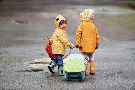 两个带着雨伞、手提箱和黄色防水斗篷和靴子的孩子雨后一起在户外散步
