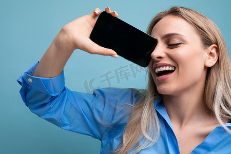 使用模型摄影照片_微笑迷人的金发女孩展示空白智能手机屏幕，带有蓝色背景网页模型