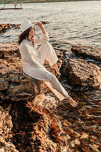 男式裤装摄影照片_一个穿着白色裤装、戴着帽子的女人站在海滩上享受大海。