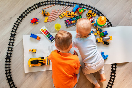 学龄前男孩在纸上的地板上画画，玩益智玩具——积木、火车、铁路、家里或托儿所的车辆。