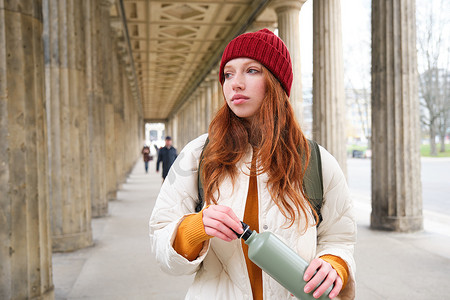 年轻的红发女孩手里拿着热水瓶，在城市里散步时给自己倒了一杯热饮。