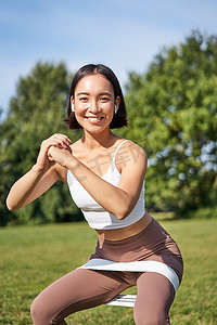 空气运动摄影照片_亚洲健身女孩在公园里蹲着，用阻力带、拉伸瑜伽绳在新鲜空气中进行锻炼训练