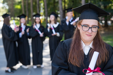 一位戴着眼镜、穿着毕业礼服的年轻白人女子在同学背景下的肖像。
