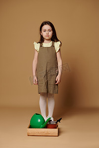 可持续资源摄影照片_清醒的小女孩站在木箱旁边，里面装着花园工具和喷壶，自信地看着镜头