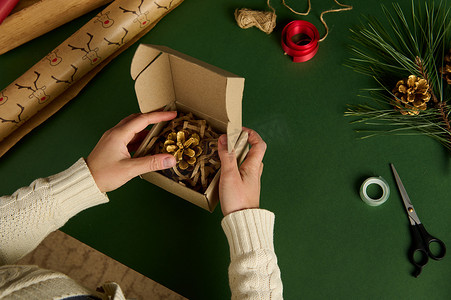 顶视图手将金色松果装入绿色背景的纸板箱中，并用装饰性包装材料