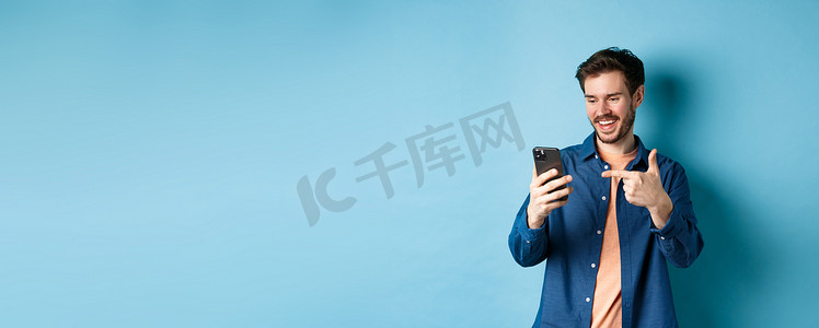 手机屏幕图片摄影照片_英俊的微笑男子站在蓝色背景上阅读手机屏幕并指着手机、查看在线促销或智能手机应用程序的图片