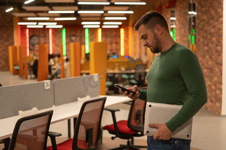 在现代联合办公空间中，白人留着胡子的男子使用智能手机并拿着笔记本电脑。