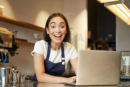 身穿咖啡馆制服的热情亚洲女孩，带笔记本电脑的咖啡师，对着镜头看起来既高兴又惊讶