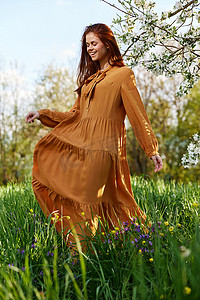 绿色树苗条摄影照片_一位快乐、苗条、甜美的女人穿着一件橙色长裙，站在一棵开花的树附近的高草丛中，幸福地微笑着，掀起了裙子的下摆，看着别处