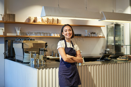 可爱的亚洲女咖啡师的肖像，咖啡馆工作人员站在柜台附近，拿着咖啡机，穿着围裙，对着镜头微笑