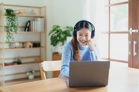 学习少女摄影照片_一名亚洲少女使用电脑、戴着耳机通过家里木桌上的视频会议在线学习的肖像