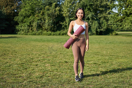春天健身摄影照片_健身女孩在公园散步、带橡胶瑜伽垫、在草坪上进行户外运动训练的肖像