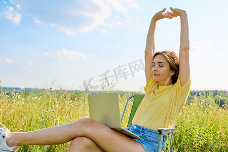 成年女性坐在折叠露营椅上，使用笔记本电脑，在一片野草丛生的草地上
