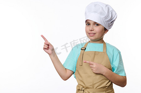 戴着厨师帽和围裙的可爱十几岁男孩指着白色背景上的促销文字的自由空间，看着相机