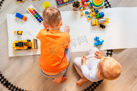 学龄前男孩在纸上的地板上画画，玩益智玩具——积木、火车、铁路、家里或托儿所的车辆。