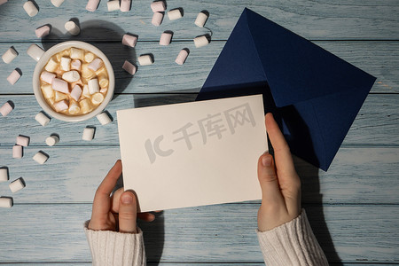 女手拿着空贺卡或邀请卡，用木制蓝色背景上的白咖啡和棉花糖模拟蓝色信封。