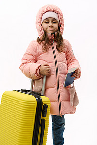 穿着粉色温暖羽绒服的可爱女婴，看着相机，摆着登机牌和黄色手提箱，白色背景