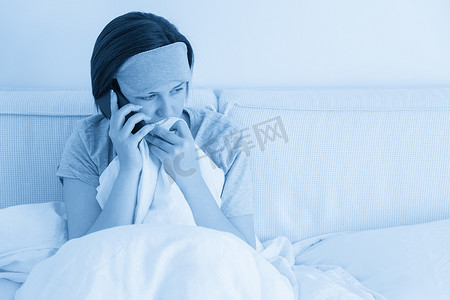 悲伤的女人戴着睡眠面罩，坐在床上，盖着羽绒被，用智能手机