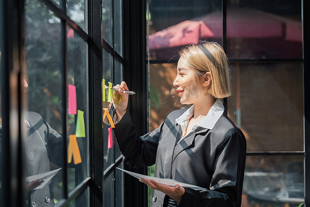 小公司的亚洲女孩女企业家在分析制定业务策略时在办公室玻璃墙上贴了贴纸