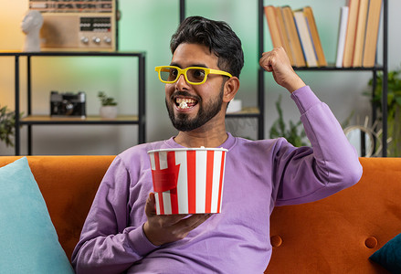 印度男子坐在沙发上吃爆米花，在家在线观看有趣的电视连续剧体育比赛