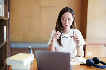 学习少女摄影照片_一名亚洲少女使用电脑和笔记本通过图书馆木桌上的视频会议在线学习的肖像