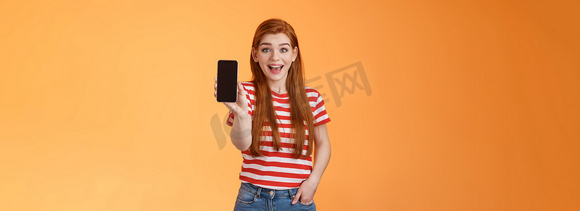 兴奋可爱的外向红发女性印象深刻的展示应用程序，持有智能手机介绍小工具功能，微笑惊讶，看起来热情像应用程序，吹牛游戏分数，橙色背景