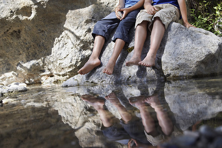 坐在河边摄影照片_两个男孩（7-9 10-12）坐在河边低处的岩石上