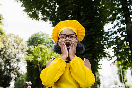 戴着厨师帽和黄色围裙制服的黑人非洲儿童厨师女孩震惊地脸颊和惊讶。