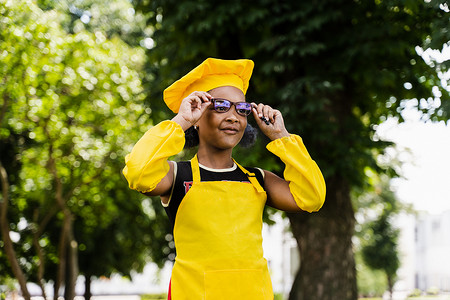 戴着厨师帽、穿着黄色围裙制服的黑人非洲儿童厨师女孩在户外微笑。