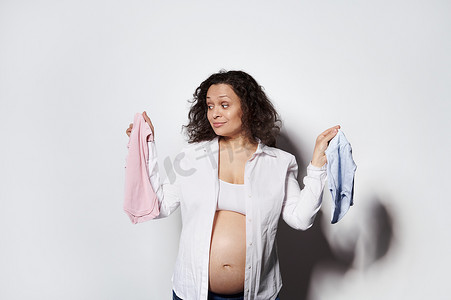 困惑的孕妇，怀孕了，拿着蓝色和粉色的新生儿衣服，猜测未来孩子的性别