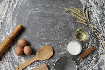 鸡蛋、面粉、橄榄油、牛奶、麦穗、灰色桌子背景上的厨房工具。