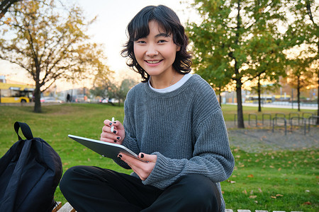 平面女孩摄影照片_年轻微笑的韩国女孩、平面设计师、艺术家用钢笔工具在数字平板电脑上绘画的肖像，坐在公园里呼吸新鲜空气，抓耳挠腮，记笔记
