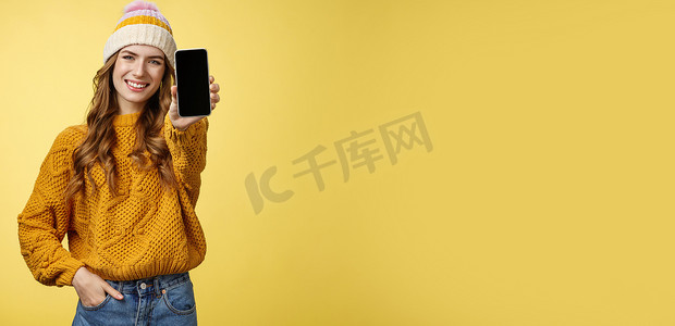 全新的摄影照片_迷人的外向微笑时尚女孩伸出手臂，向您展示全新的智能手机，显示满意的咨询朋友使用应用程序编辑照片手机，黄色背景放置什么过滤器