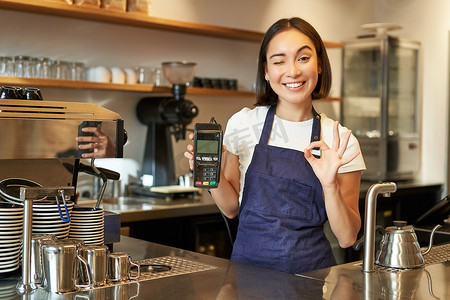 微笑的亚洲咖啡师女孩，穿围裙，展示信用卡机处理付款，建议非接触式付款，站在咖啡店