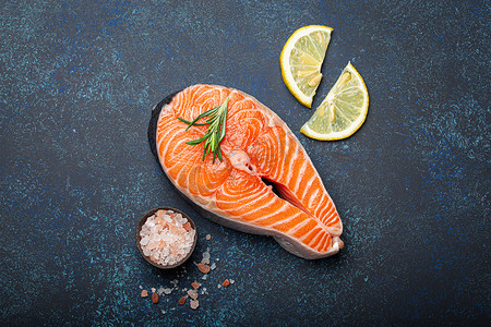 深蓝色质朴石背景上的新鲜生鲑鱼片牛排，配迷迭香，配以玫瑰盐和柠檬顶景，健康均衡的饮食和营养