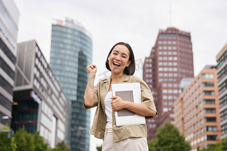 市中心快乐的亚洲女孩，表现出“是”的万岁姿态，站着笔记本电脑庆祝