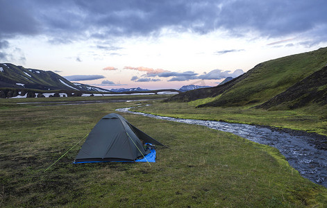 小绿色帐篷矗立在溪岸露营地 Strutur 的绿草上，靠近 f210 路、积雪覆盖的山丘、午夜粉红色的夕阳天空，冰岛