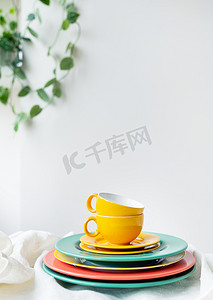 黄色转轮摄影照片_白色的桌子上铺着亚麻桌布，上面放着空的黄色杯子和色彩鲜艳的盘子，背景墙上有一个花盆。