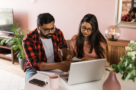 年轻的千禧一代印度丈夫和妻子做家庭文书工作、会计工作并审查纸质账单、笔记本电脑收据、使用在线计算器并在互联网上支付抵押贷款租金 — 经济危机概念