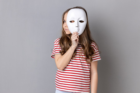 诚实的摄影照片_可爱的小女孩拿着白色面具，表情严肃地看着别处。