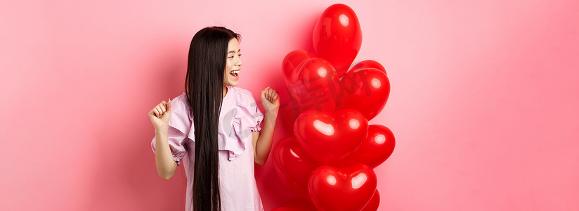 长发的亚洲少女，从情人节浪漫的礼物中欢呼，看着标志，开心地微笑，从情人礼物心形气球附近的喜悦中跳起来，粉红色的背景