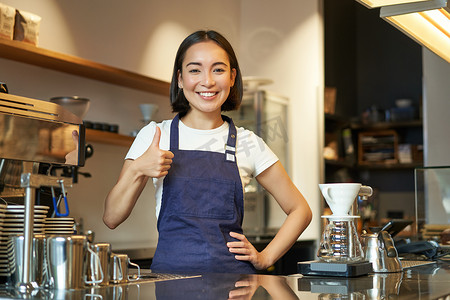 微笑的亚洲咖啡师，穿着咖啡馆制服的女孩，表现出好的迹象，推荐他们的咖啡，批准新口味，站在围裙