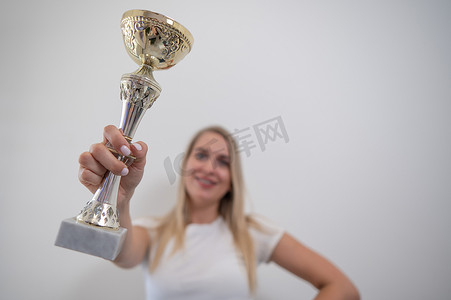 奖杯墙摄影照片_金发碧眼的女人微笑着，拿着一个金色的奖杯靠在白色的墙上。