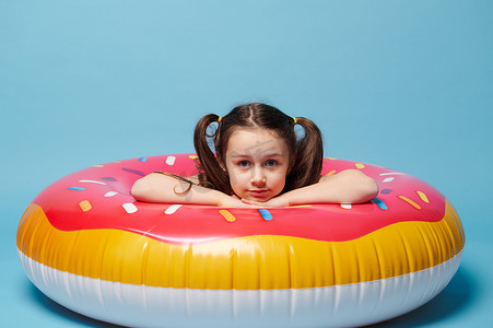 儿童游泳圈摄影照片_穿着泳衣的小女孩，在游泳圈内 — 一个彩色的粉红色甜甜圈，对着镜头微笑，孤立的蓝色背景。