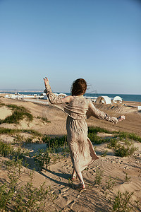夏天大海沙滩背景摄影照片_一位身着米色连衣裙的优雅女子在沙滩上行走，背景是远处可见的大海，背对着镜头