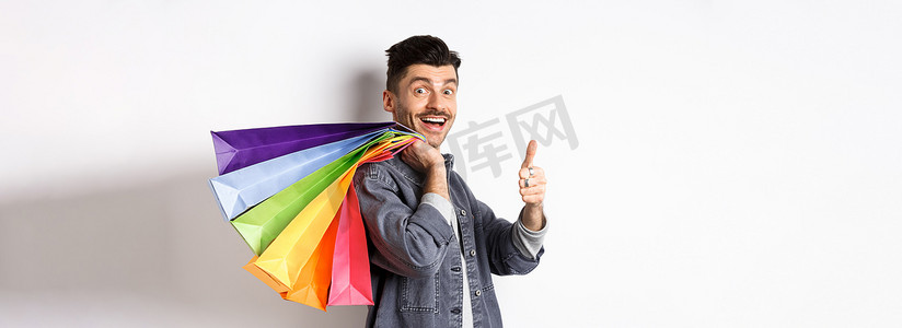 优惠商品摄影照片_兴奋的男性购物者肩上扛着购物袋，竖起大拇指，站在白色背景上推荐商店里的优惠商品