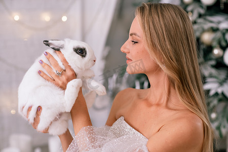 拿着 2023 年白兔象征的女人。一位美丽的年轻金发女郎的特写镜头，她抱着一只穿着闪闪发光的裙子的兔子。